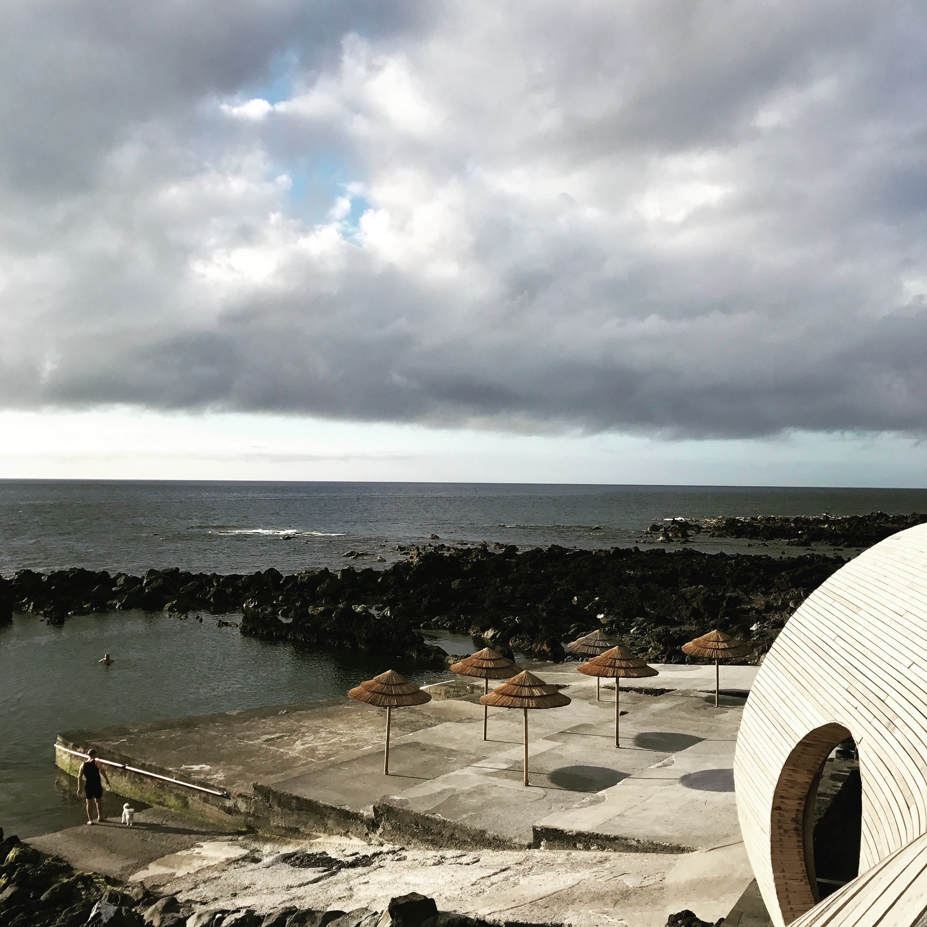 Rocky ocean pools and straw parasols beside a curved white building.