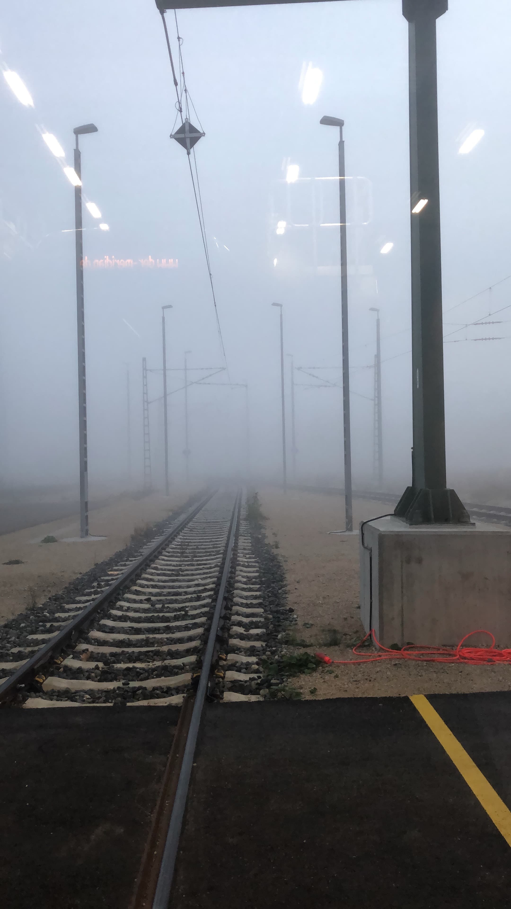 Railway tracks and overhead wires disappear into thick fog, seen through a train window.